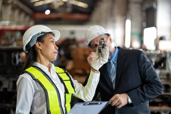 Una ingeniera con casco y chaleco de seguridad, guantes blancos, y un hombre representan un control de calidad bajo la ISO 45001.