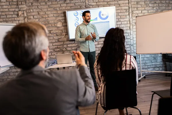 Un presentador sonriente explica gráficos en una pantalla, ideal para un curso o Diplomado en Normas ISO.